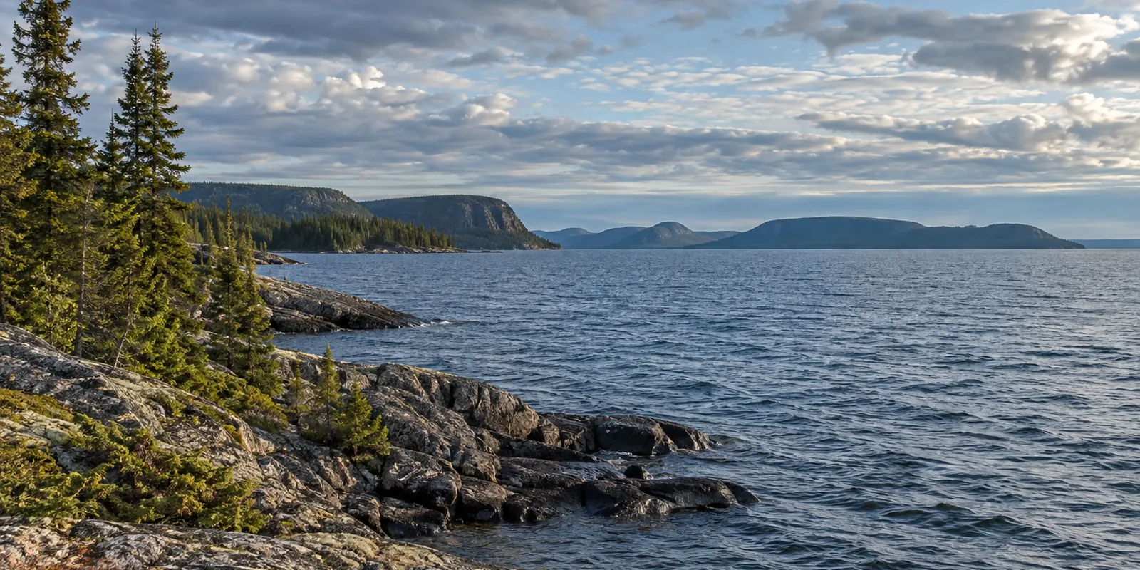 A cold blue northern lake with a rocky spruce shoreline and low rugged hills beneath broken clouds.